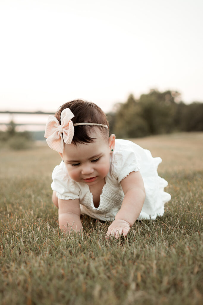 Baby crawling in grass wearing white dress and pink bow posing for a Huntsville Photographer family photoshoot