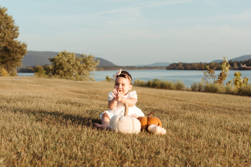 Baby sitting grass posing with pumpkins wearing white dress and pink bow posing for a Huntsville Photographer family photoshoot